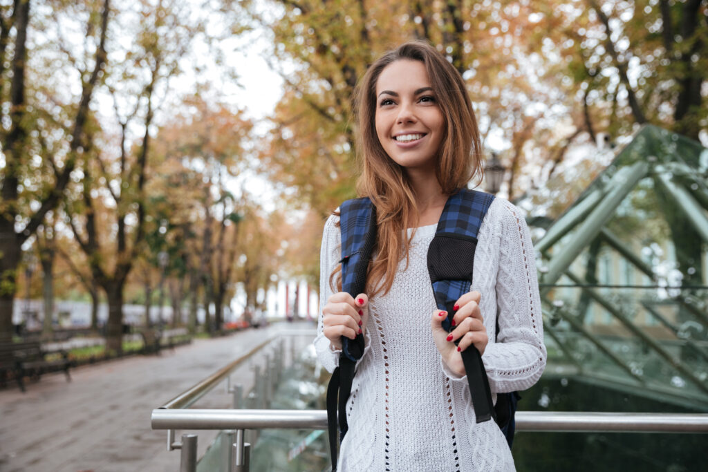 Cheerful charming young woman with backpack walking in park